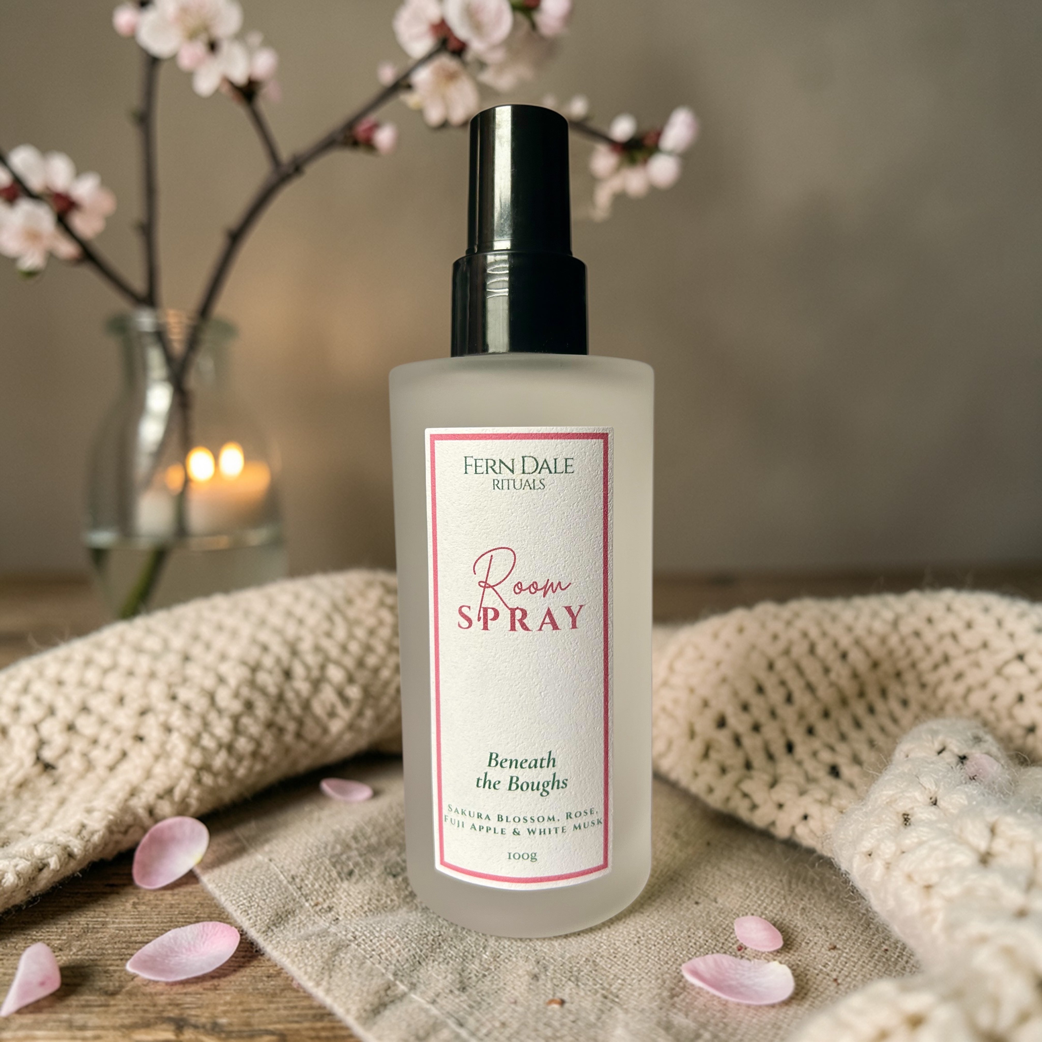 Beneath the Boughs Room Spray in a frosted bottle with a black cap, surrounded by pink petals and cream knitted fabric.