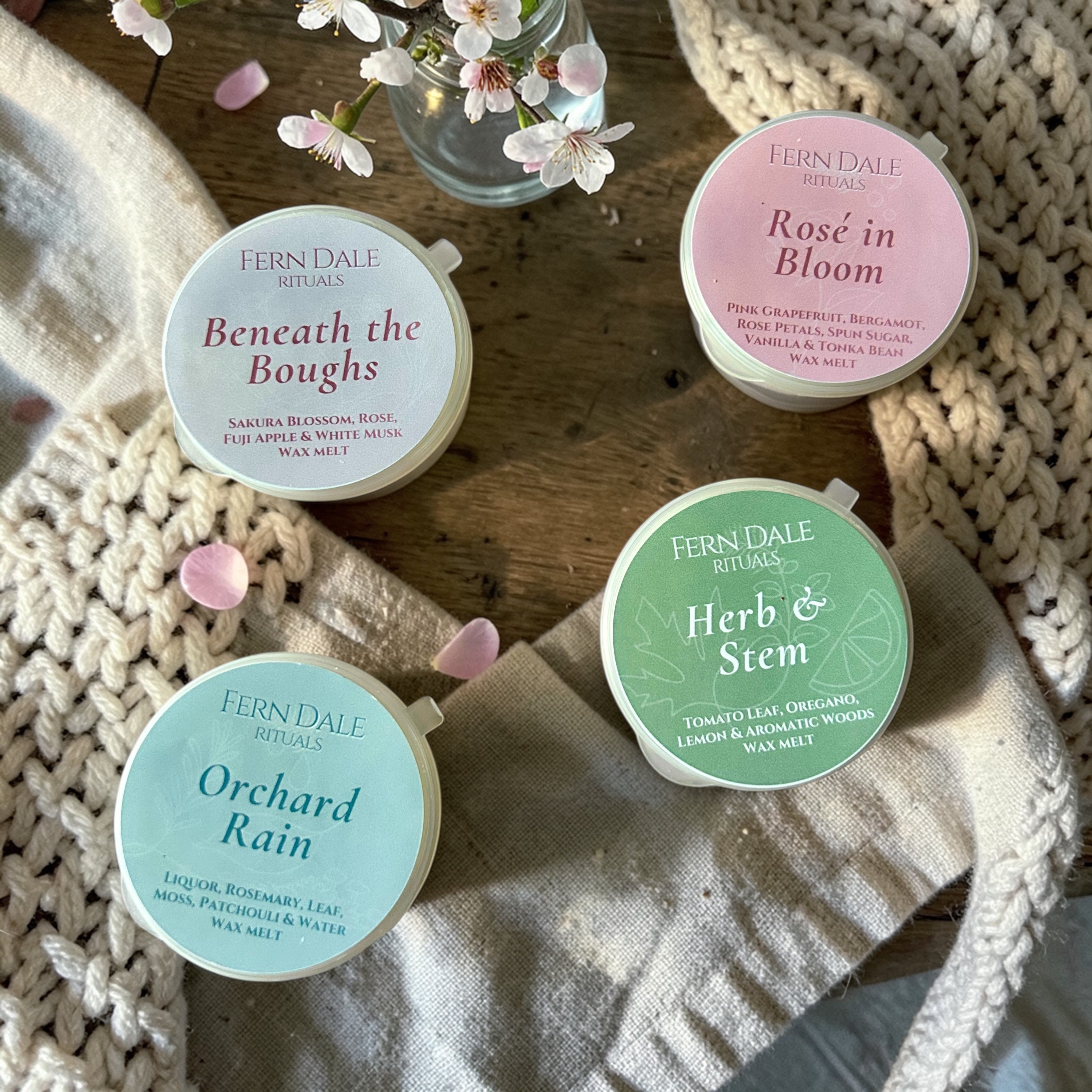 Beneath the Boughs Wax Melt Collection in four round tubs with pastel labels, beside blossom and cream fabric.