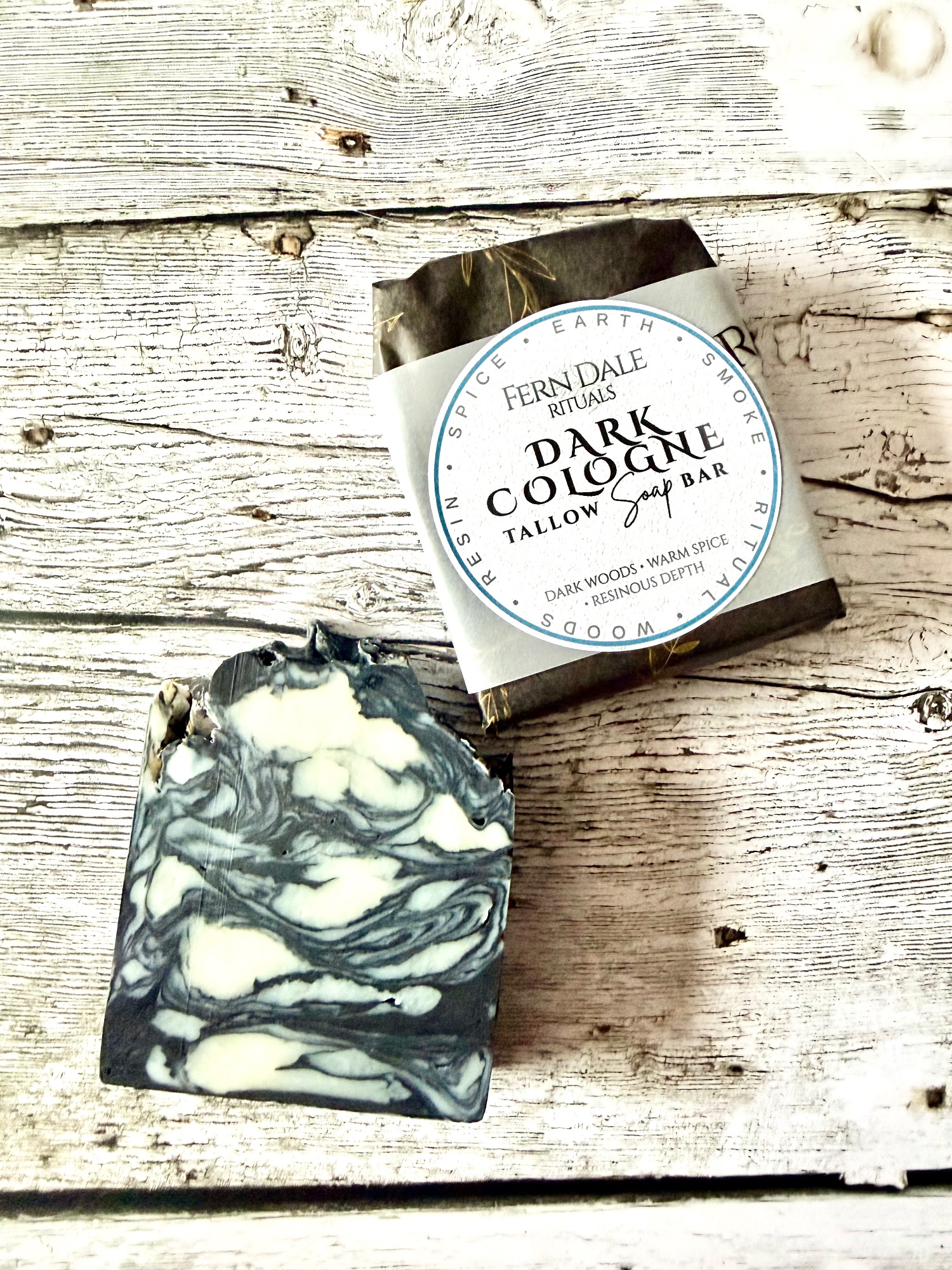 Dark Cologne Tallow Soap with dark grey and cream swirls beside a wrapped bar on a whitewashed wooden surface.