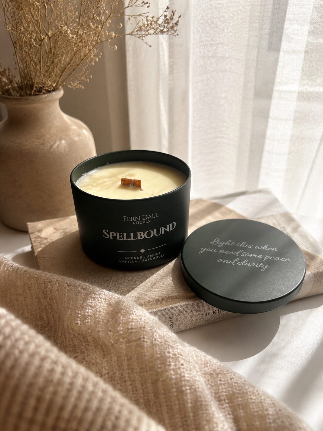 Engraved Candles in a dark green tin with a wooden wick and an engraved lid beside a vase of dried flowers.