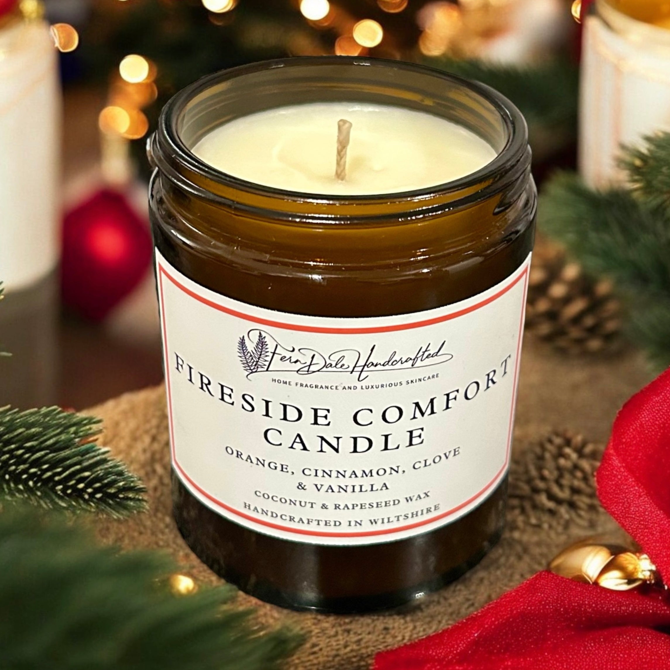 Fireside Comfort Candle unlit in an amber glass jar with a white label, surrounded by greenery and red ribbon.